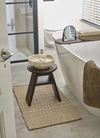 Badteppich in Beige mit strukturierter Oberfläche und dunklem Holzhocker neben einer weißen Badewanne
