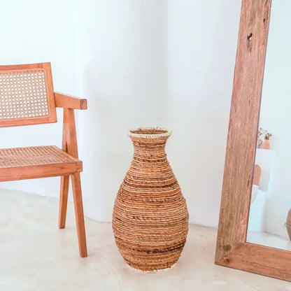 Geflochtene Bodenvase Tumbak aus Bananenfaser neben Holzstuhl im Natur-Look.